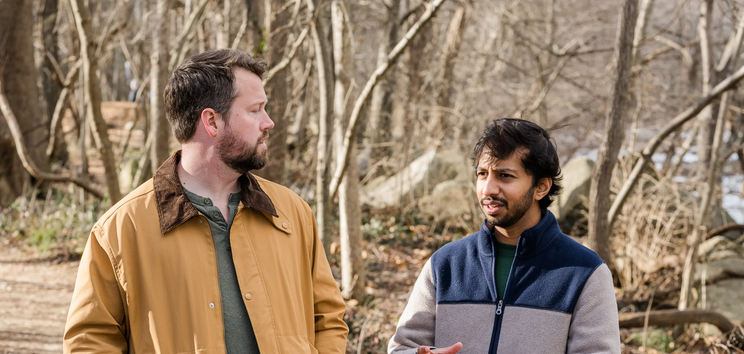 Two men walking and talking on a bare-branched trail in winter light